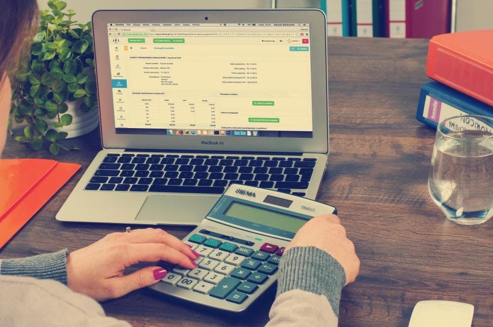 Pessoa usando calculadora ao lado de um notebook com planilha aberta